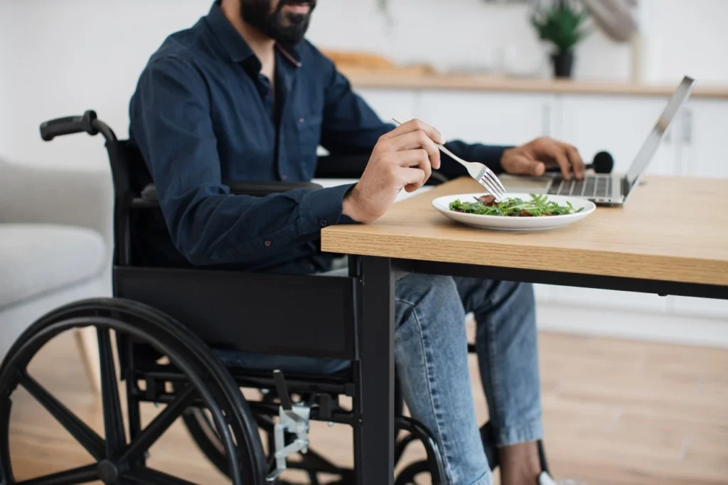 Man in wheelchair eating a meal at table while using laptop